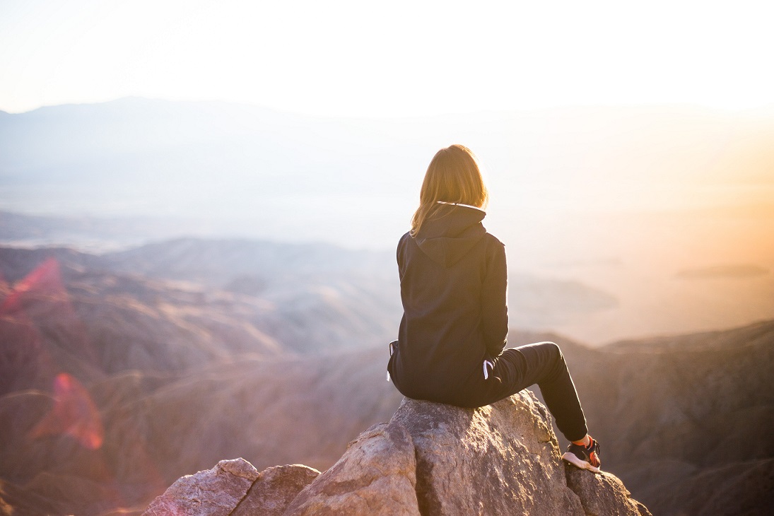 Photo of the back of a woman, sitting on a rock overlooking a canyon at sunset.